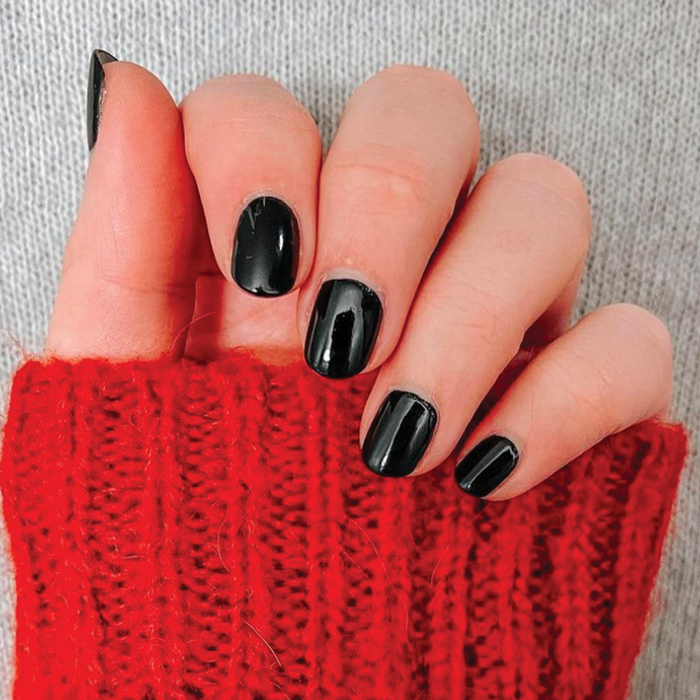 Hand with short nails painted shiny black polish wearing a vibrant red knitted sweater sleeve against gray fabric background