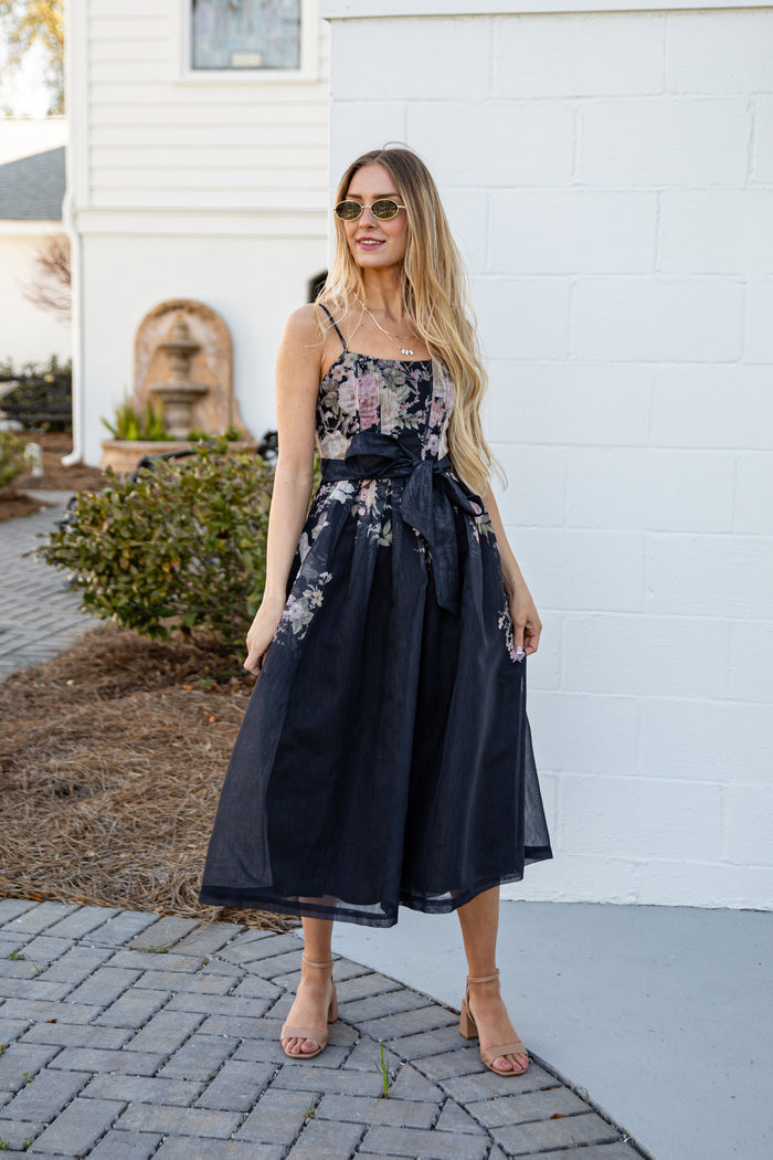 Woman wearing a floral dress with a black overlay standing in front of a white wall.