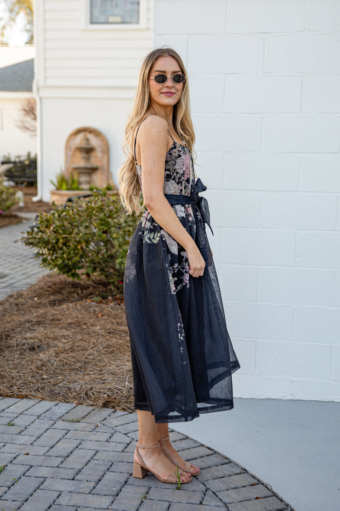 Woman in a floral dress standing against a white brick wall.