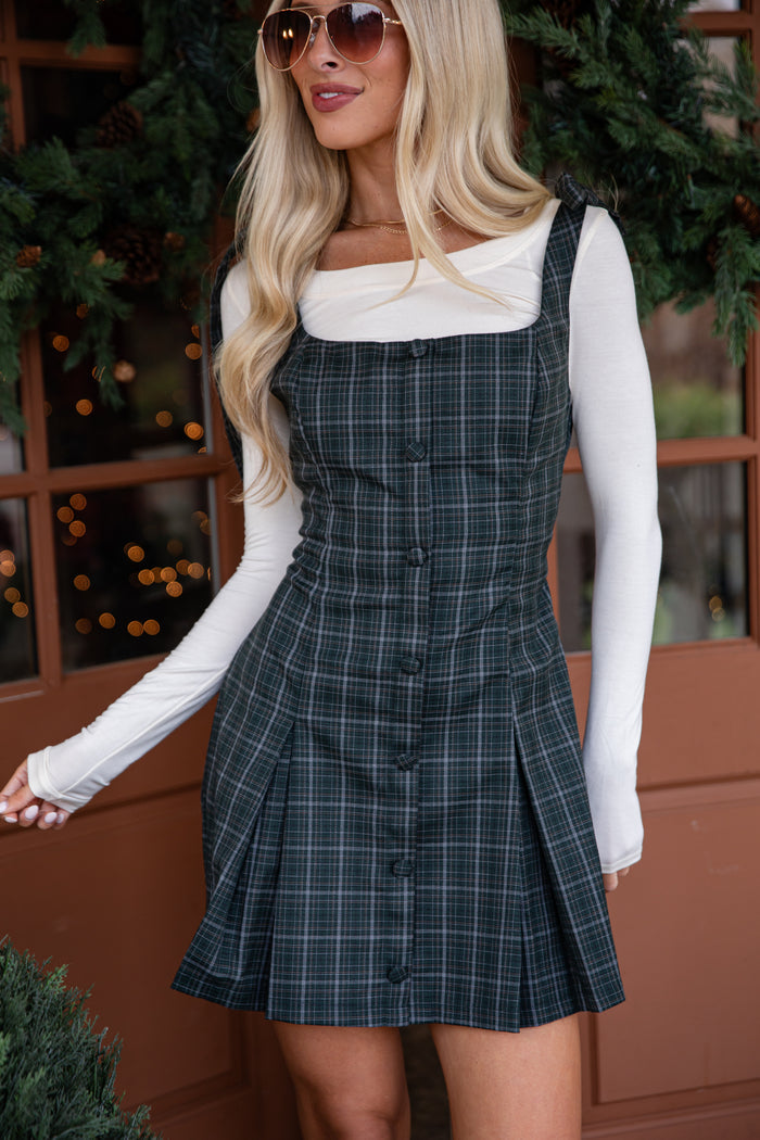 Woman wearing a plaid dress with a white long-sleeve shirt and sunglasses, standing in front of a decorated wall.