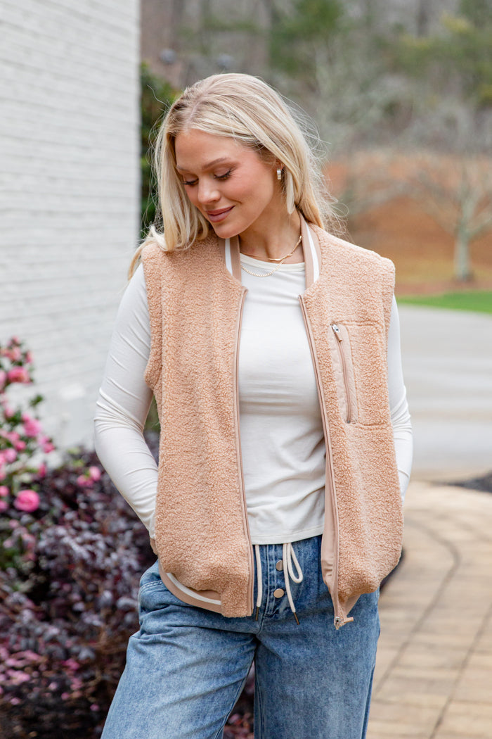 Woman wearing a beige vest over a white shirt and blue jeans, standing outdoors.
