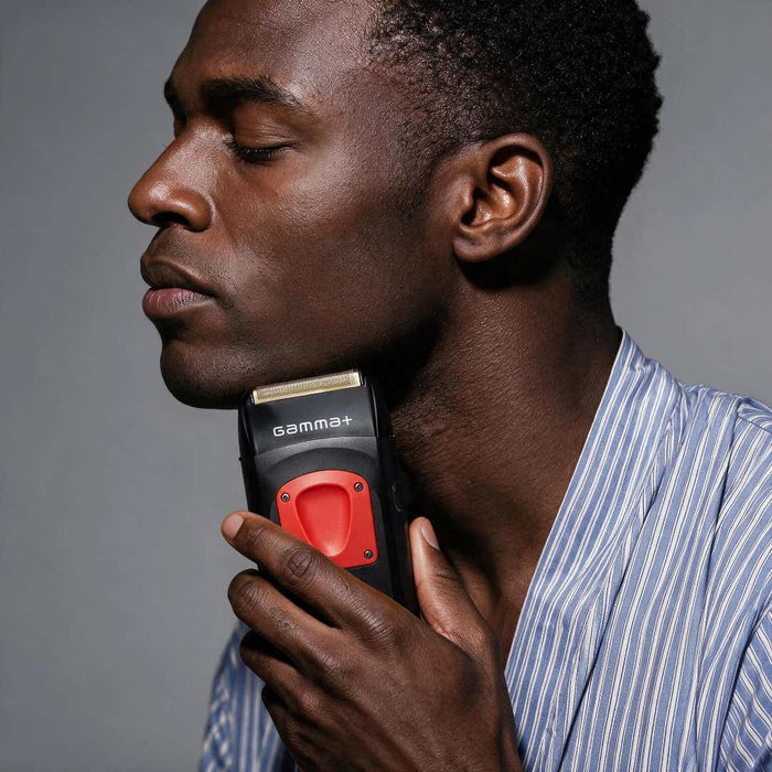 A close-up of a man with closed eyes holding a black and red Gamma+ electric trimmer against his neck, wearing a blue and white striped shirt against a solid gray background.