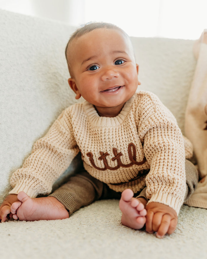 Smiling baby wearing Viverano Organics beige knitted top made from organic cotton sitting on a couch