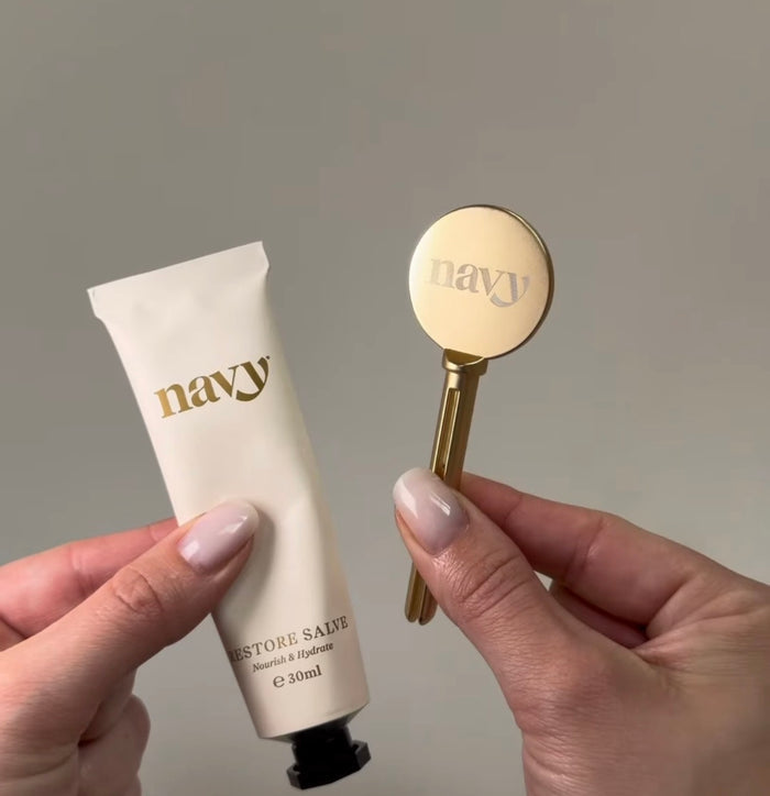 Hand holding a tube of 'navy' restore salve and a applicator against a neutral background