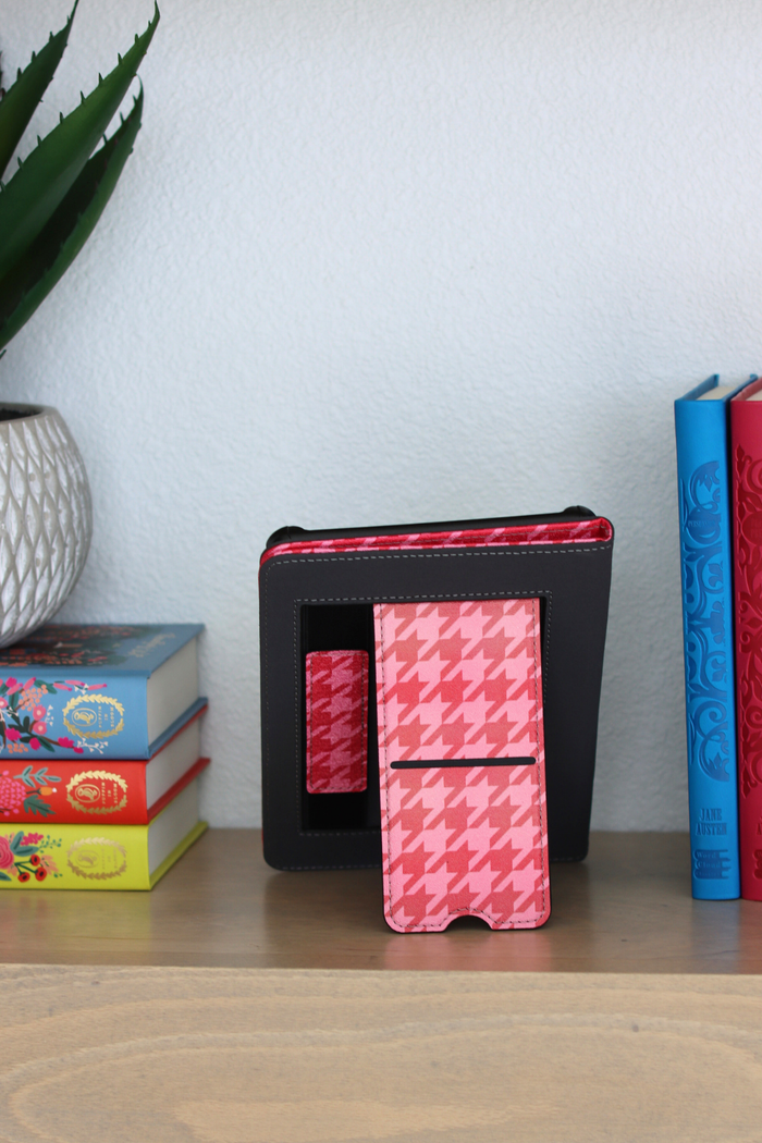 Pink and red kindle on a shelf with books and a plant in the background