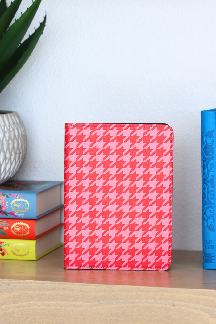 Pink houndstooth patterned kindle on a wooden surface with books and a plant in the background.