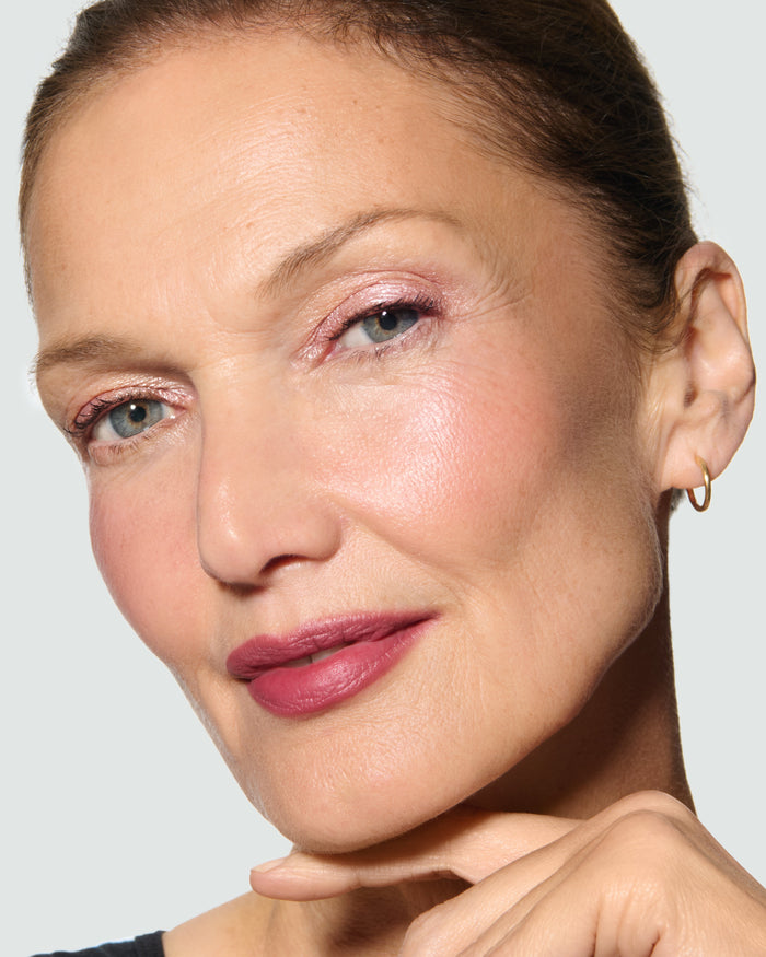 Close-up of a model's face against a neutral background, wearing Rosalie Liquid Lurex Liquid Eyeshadow, a warm, peachy rose.
