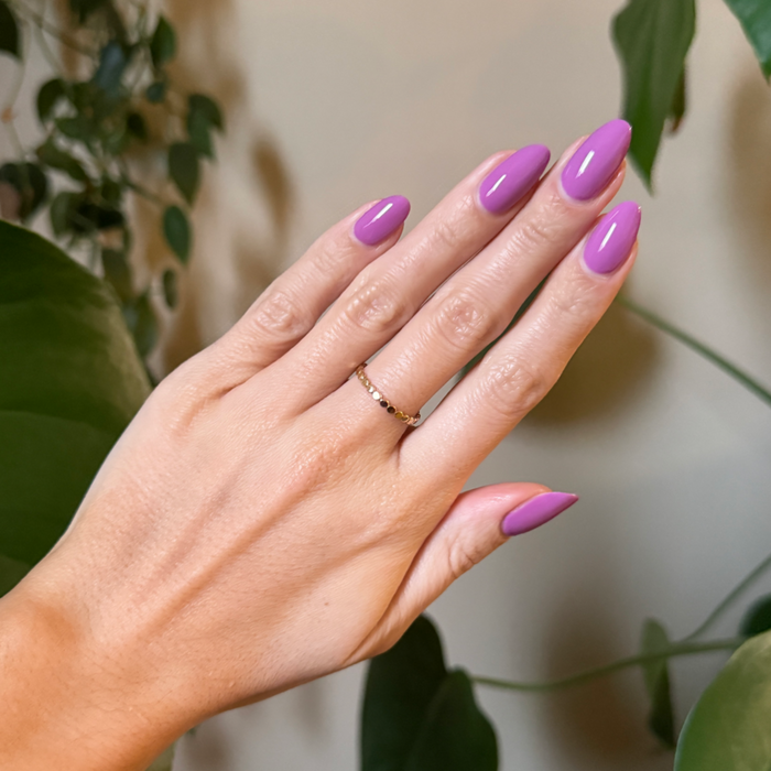 Hand with almond-shaped nails polished in bright floral purple pink using Petal - Gel against leafy natural backdrop