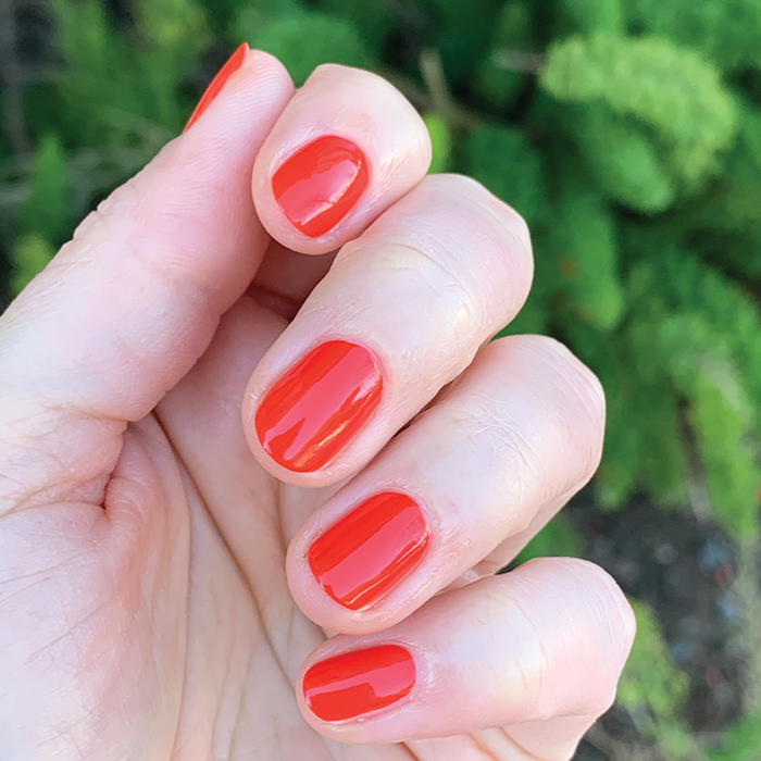Hand with neatly applied Lava nail polish in vibrant spicy orange red against a natural green outdoor background