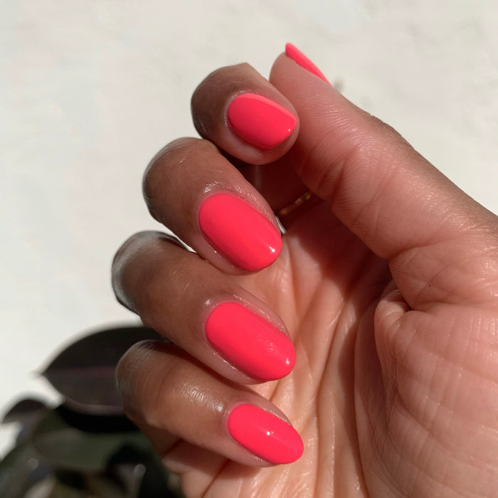 Hand with oval nails coated in bright coral-pink gel polish against a soft off-white background