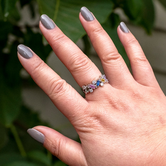 Hand with nails painted in AW warm toned grey showing a smooth manicure with a colorful gemstone ring against a leafy background