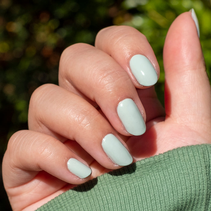 Hand with nails painted in soft sage green KMC gel polish showing a smooth, natural manicure in bright sunlight