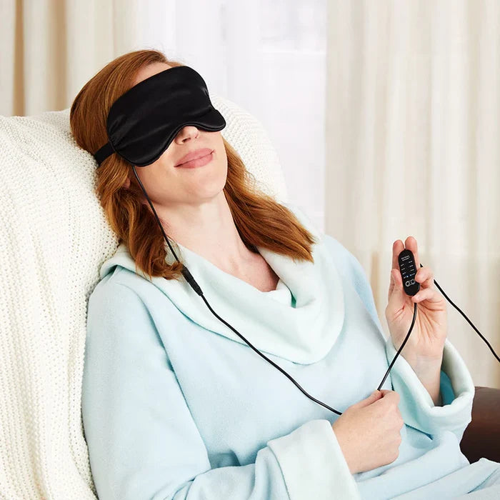 A woman relaxing in a chair while wearing a black heated sleep mask and holding the connected remote control.